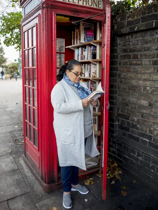 Seorang wanita membaca buku di dekat kotak telepon merah yang telah berubah menjadi perpustakaan pertukaran buku di Lewisham Way, London (21/10). Kotak telepon ini diubah menjadi perpustakaan, kios, hingga tempat reparasi ponsel. (AFP Photo/Tolga Akmen)