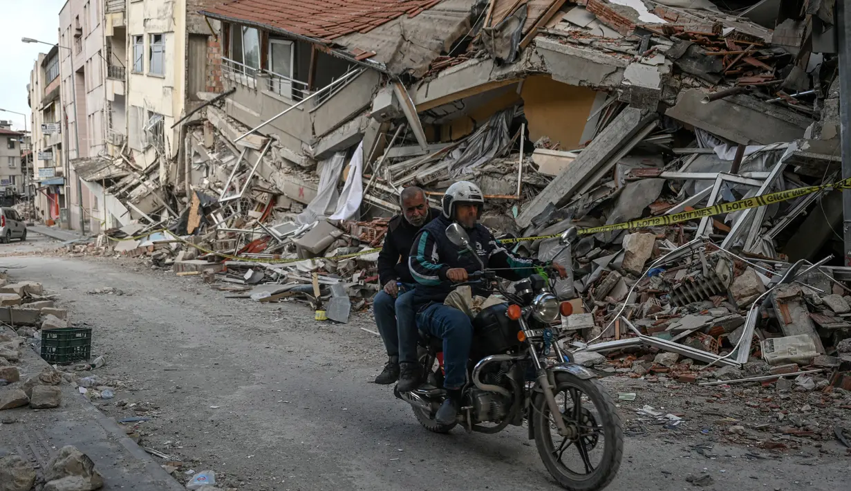 <p>Warga mengendarai sepeda motor di jalan di sebelah puing-puing bangunan yang runtuh di Hatay pada 6 Maret 2023, satu bulan setelah gempa besar melanda Turki tenggara. Gempa bumi besar berkekuatan magnitudo 7,8 mengguncang sebagian besar Turki dan sebagian Suriah pada 6 Februari 2023, menewaskan lebih dari 50.000 orang di kedua negara, dengan sekitar 46.000 di pihak Turki. (OZAN KOSE / AFP)</p>