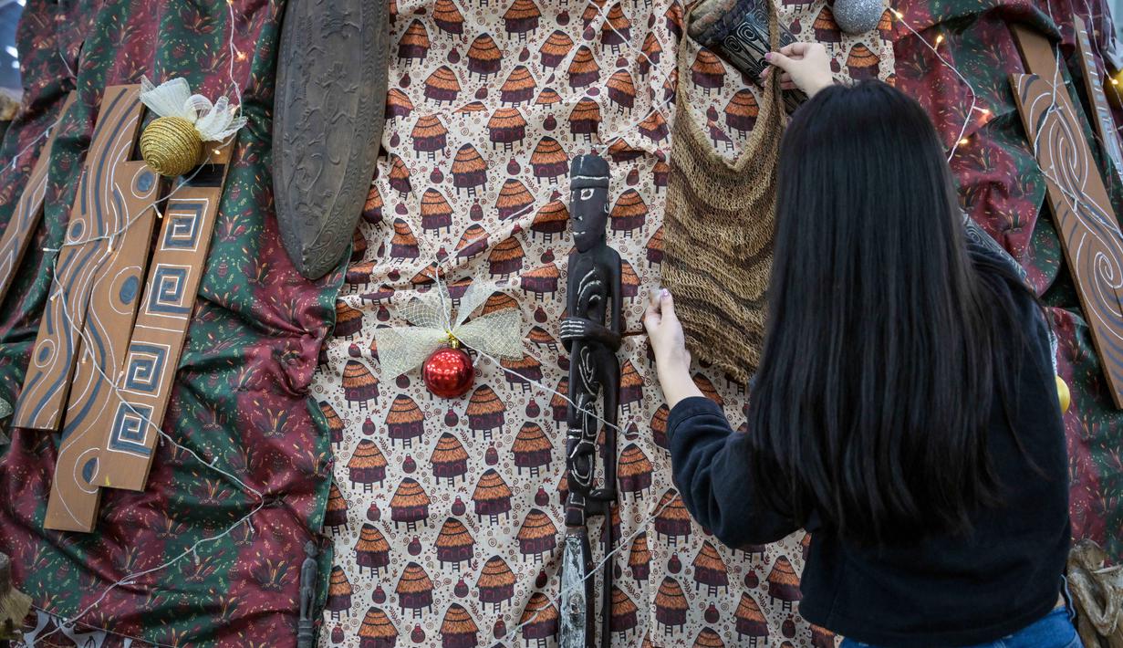Instalasi dekorasi Pohon Natal ini berdiri setinggi tujuh meter dengan diameter 3,3 meter dan dibuat dari rangka kayu serta kain bermotif khas Papua. Tampak dalam foto, salah satu mahasiswa memasang sentuhan akhir pada instalasi dekorasi berbentuk pohon Natal dengan motif budaya Papua di Perpustakaan Petra Christian University (PCU), Surabaya, Jawa Timur, Kamis 18 Desember 2025. (JUNI KRISWANTO/AFP)