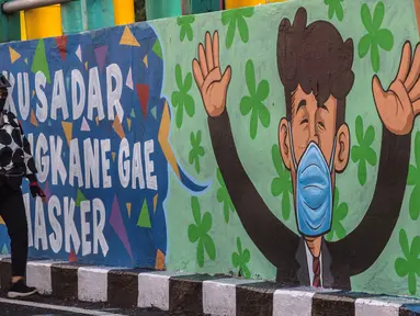 Seorang perempuan melewati mural yang mengajak orang untuk memakai masker di tengah pandemi Covid-19 di Surabaya, Minggu (25/10/2020). Mural di sepanjang dinding itu sebagai sarana imbauan kepada masyarakat untuk menerapkan protokol kesehatan pencegahan penularan COVID-19. (Juni Kriswanto/AFP)