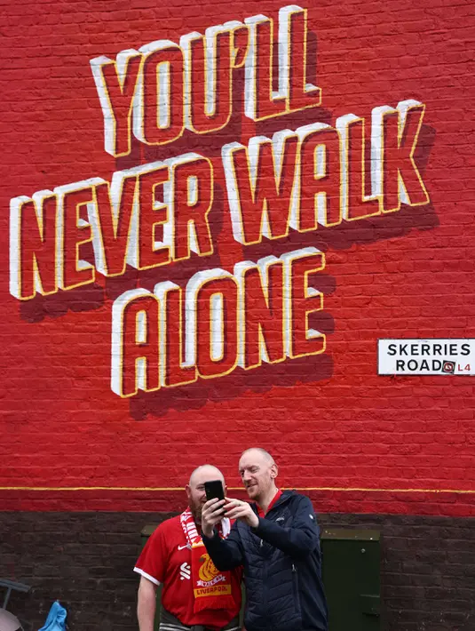 Dua pendukung Liverpool berfoto di depan tembok yang bertuliskan "You'll Never Walk Alone" sebelum laga lanjutan Liga Inggris 2024/2025 melawan Brentford di Anfield, Liverpool, Inggris, Minggu (25/08/2024) malam WIB. (AFP/Darren Stapless)
