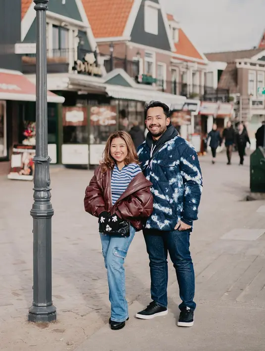 Kiky Saputri dan suaminya berpose dengan outfit bernuansa biru yang senada. Kiky memilih mengenakan kaus garis-garis biru-putih yang ditumpuknya dengan puffer jacket berwarna merah bata, dipadu dengan celana panjang jeans. Sedangkan suaminya memilih mengenakan hoodie hitam yang ditumpuknya lagi dengan oversized jacket tie dye bernuansa biru-putih, dipadu dengan celana panjang jeans. Foto: Instagram.