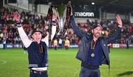 Aktor Hollywood, Ryan Reynolds (kanan) dan pemilik klub Wrexham, Rob McElhenney merayakan kesuksesan dengan mengangkat trofi setelah klub mereka berhasil promosi ke Liga Dua Inggris. Wrexham menang atas Boreham Wood dengan skor 3-1 pada laga English National League di Racecourse Ground Stadium, Wrexham, Wales, Sabtu (22/04/2023). (AFP/Oli Scarff)