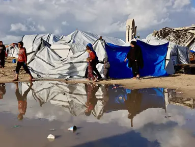 Warga Palestina yang mengungsi berjalan melewati genangan air di dekat tenda-tenda penampungan saat wilayah tersebut dilanda hujan dan kondisi musim dingin yang dingin, di Kota Gaza pada 28 Desember 2025. Cuaca ekstrem serta musim dingin yang ekstrem di Jalur Gaza semakin memperparah krisis kemanusiaan di tengah keterbatasan tempat berlindung yang aman. (Omar AL-QATTAA/AFP)