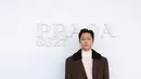 Lee Do-Hyun tampil dengan setelan jas cokelat berkerah bulu domba hitam, dipadukan turtleneck krem dan tas kulit hitam. [dok. Prada]