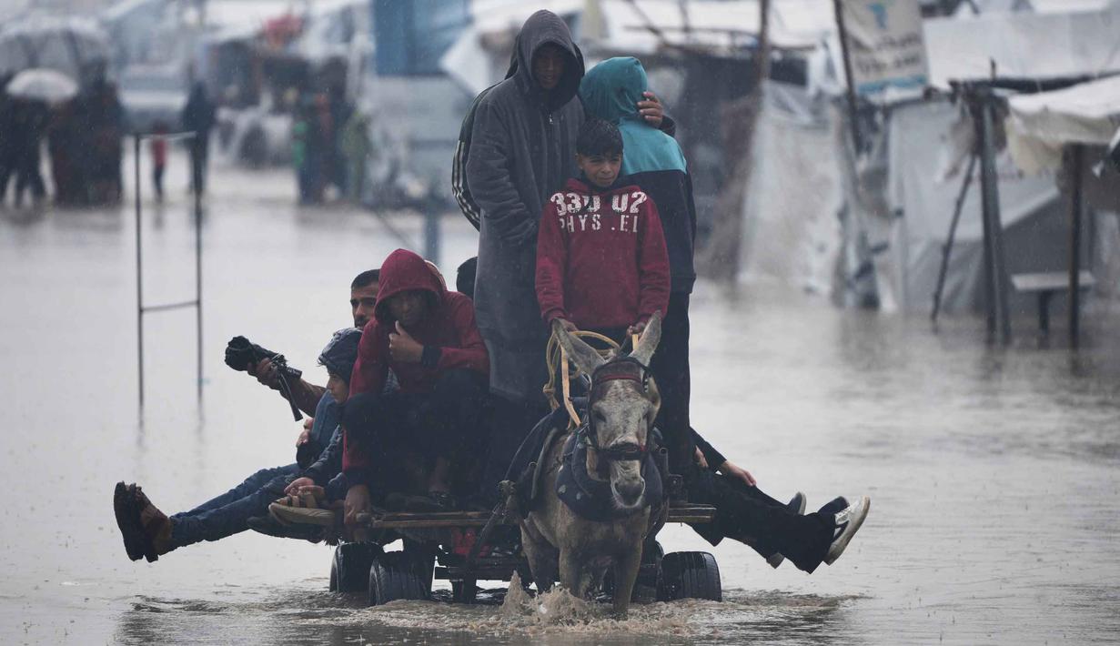 Puluhan hingga ratusan tenda pengungsi Palestina di wilayah tersebut terendam banjir. Bahkan, sebagian besar tenda pengungsi dilaporkan tidak lagi layak huni. Tampak dalam foto, warga Palestina menyeberangi jalan yang tergenang banjir setelah hujan lebat di Khan Younis, Jalur Gaza selatan, Kamis 11 Desember 2025. (AP Photo/Abdel Kareem Hana)