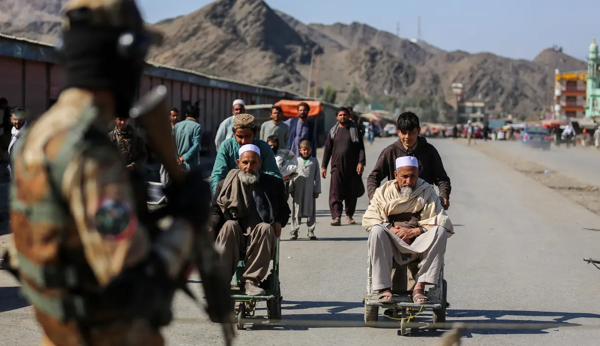 Seorang personel keamanan Taliban berjaga-jaga ketika anak laki-laki Afghanistan membantu pria lanjut usia di kursi roda setelah insiden baku tembak antara pasukan perbatasan Afghanistan dan Pakistan di dekat perbatasan Torkham yang melintasi antara Afghanistan dan Pakistan, di provinsi Nangarhar pada 20 Februari 2023. Penyeberangan utama di perbatasan Afghanistan-Pakistan tetap ditutup pada Selasa, 21 Februari 2023, untuk hari ketiga berturut-turut, kata para pejabat. (Shafiullah KAKAR / AFP)