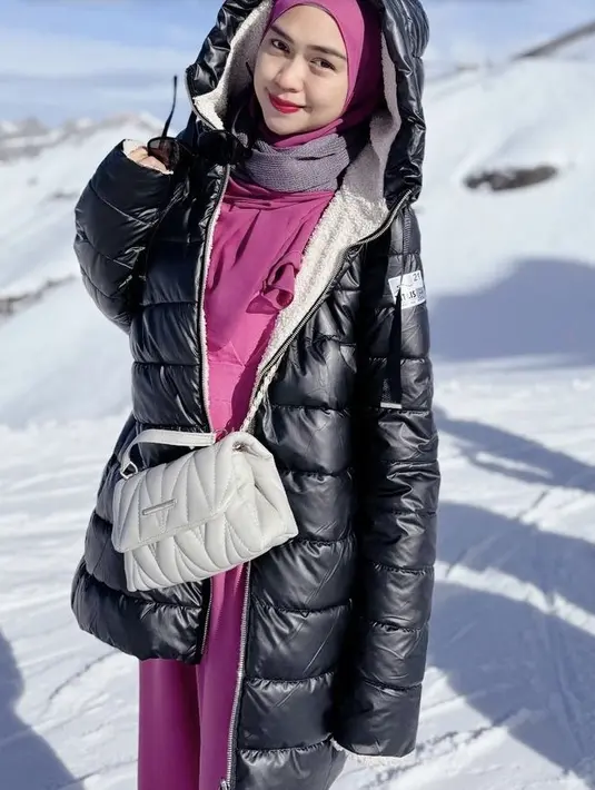 Ria Ricis mengenakan jaket padding hitam dipadukan gamis dan kerudung fuschia. Sambil membawa sling bag putih. [@riaricis1795]