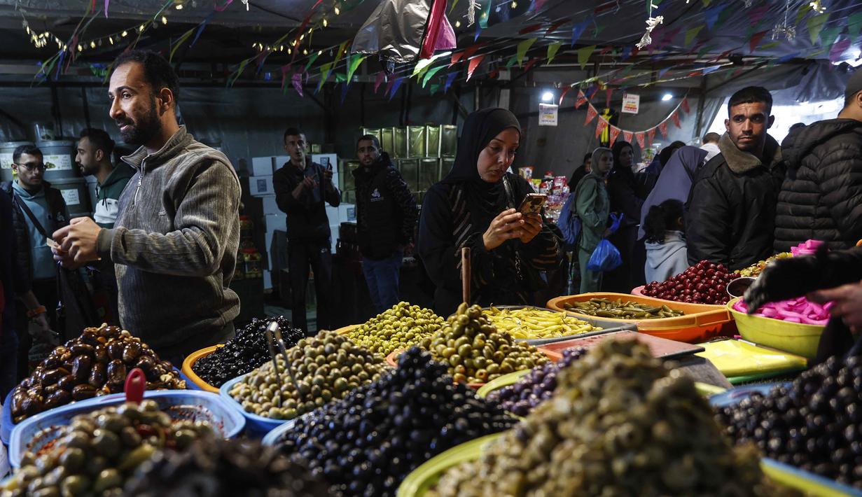 Sejumlah kios berjejer rapi, menawarkan beragam kebutuhan seperti acar, kurma, dan rempah-rempah yang menjadi incaran para pembeli. Tampak dalam foto, seorang wanita Palestina berdiri di depan sebuah toko yang menjual zaitun di pasar Zawiya, Kota Gaza, pada Rabu 18 Februari 2026. (Omar AL-QATTAA/AFP)