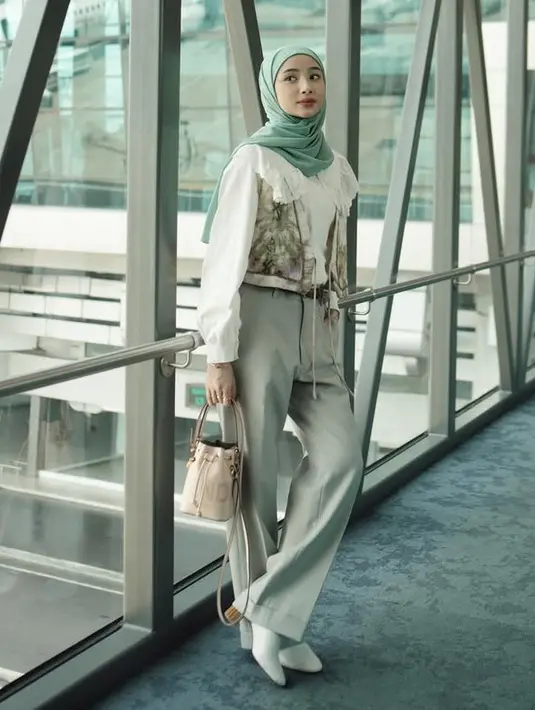 Airport outfit mengenakan kemeja putih dengan kerah collar lace, Anselma memadukannya dengan outer vest tie dye serta kerudung hijau. Ia mengenakan bawahan celana panjang abu-abu serta heels tertutup depan warna putih. Sambil membawa bucket bag. [@ansellmaputri]