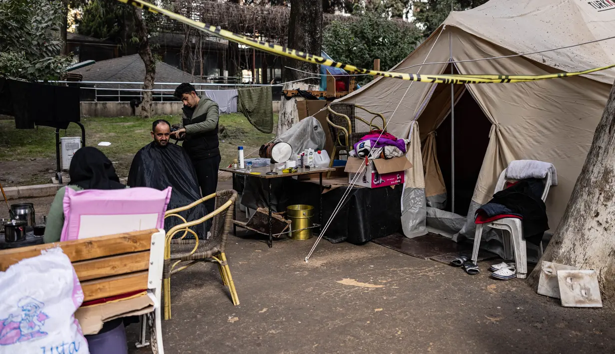 Mohammed al-Hamo (kanan) memotong rambut ayahnya Khaled di depan tenda mereka di kamp darurat pengungsi korban gempa Turki dan Suriah di kota Antakya pada 19 Februari 2023. Hamo yang kini berusia 18 tahun awalnya belajar memotong rambut untuk mendapatkan penghasilan sampingan di waktu luangnya. (Sameer Al-DOUMY / AFP)