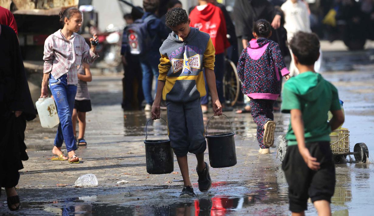 Seorang pemuda Palestina membawa ember-ember air yang diisi di titik pengambilan air di kamp pengungsi Nuseirat, yang terletak di Jalur Gaza tengah, pada 2 November 2025. Selain itu, sekitar 650.000 anak perlu segera kembali bersekolah. Hal tersebut dinyatakan Tess Ingram pada Minggu 2 November 2025. (EYAD BABA/AFP)