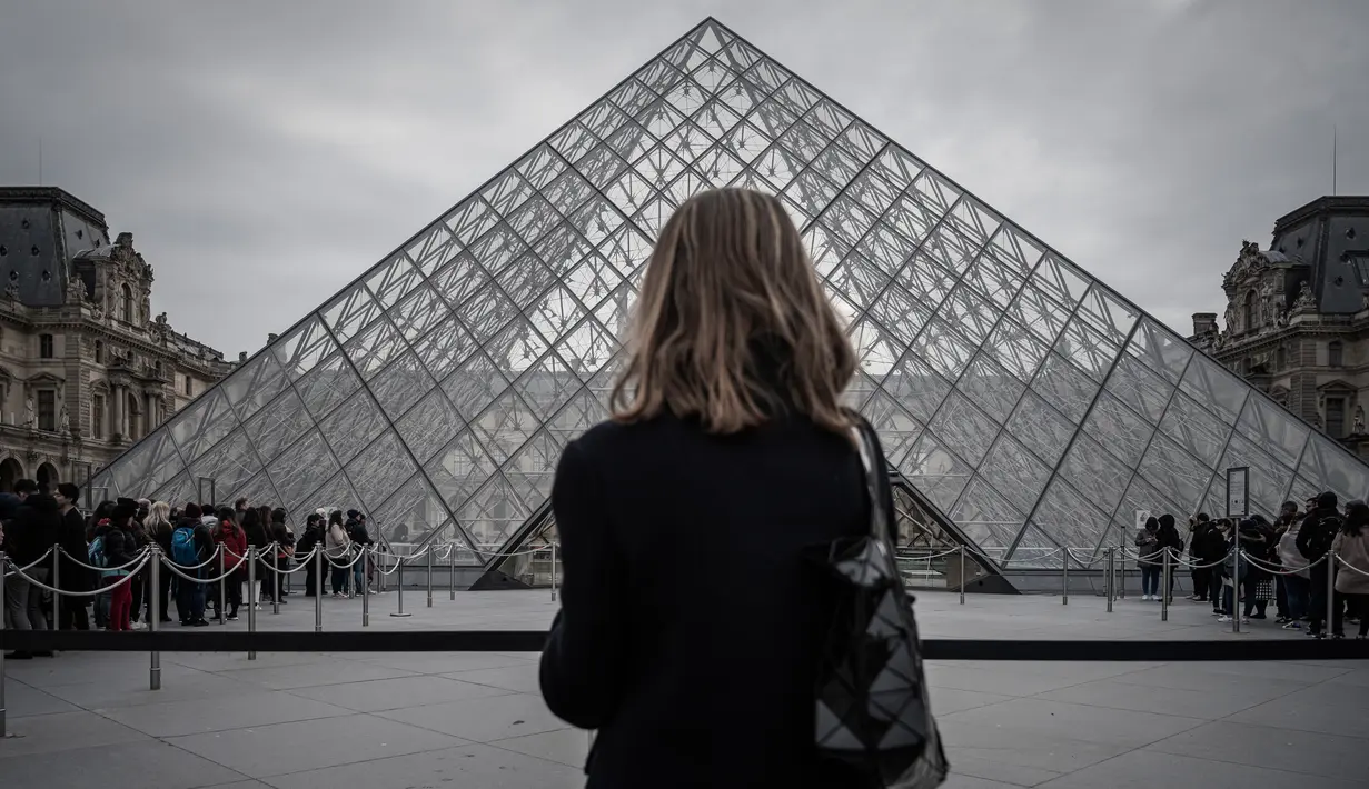 Seorang perempuan berdiri di depan Pyramid, pintu masuk utama menuju museum Louvre, di Paris, Rabu (4/3/2020). Louvre di Paris, museum yang paling banyak dikunjungi di dunia, kembali dibuka setelah pegawai yang khawatir dengan penularan virus corona setuju kembali masuk kerja. (Philippe LOPEZ/AFP)