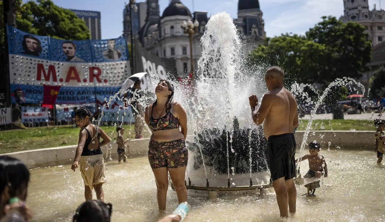 Orang-orang mendinginkan diri di air mancur, denggan latar belakang protes yang diselenggarakan oleh berbagai organisasi sosial berlangsung, di Buenos Aires, Argentina, Jumat (3/3/2023). Menurut Badan Meteorologi Nasional, Buenos Aires tetap dalam status siaga merah untuk suhu ekstrem. (AP Photo/Rodrigo Abd)