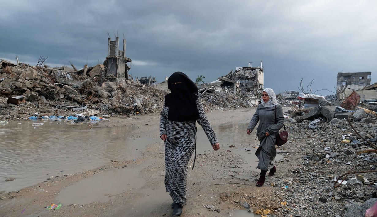 Hujan lebat dan cuaca dingin yang melanda di Jalur Gaza menyebabkan banjir dan memperumit situasi kemanusiaan bagi warga Palestina di Gaza. Tampak dalam foto, warga Palestina berjalan melewati reruntuhan di tengah cuaca buruk Kota Gaza, Kamis 11 Desember 2025. (AP Photo/Jehad Alshrafi)