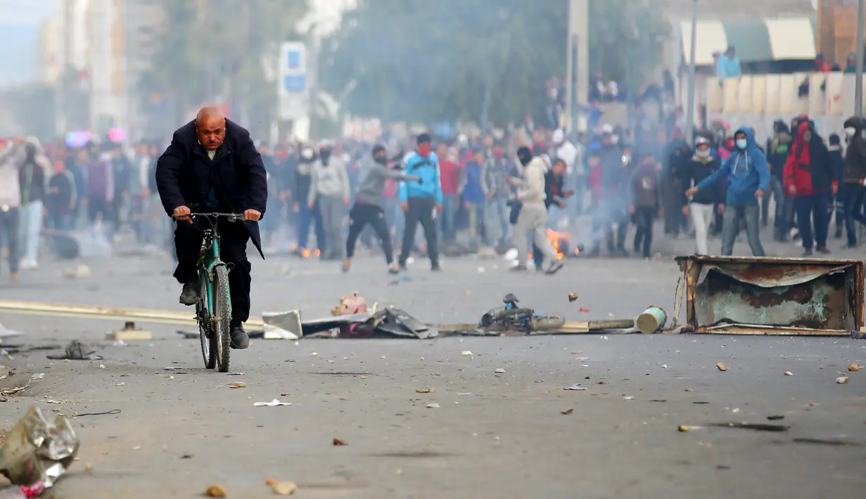 Seorang pria mengendarai sepeda saat pengunjuk rasa melemparkan batu ke arah polisi di luar kantor pemerintah setempat di Kasserine, Tunisia, Kamis (21/1). Mereka menuntut solusi dari masalah pengangguran yang meliputi negaranya. (REUTERS/Amine Ben Aziza)