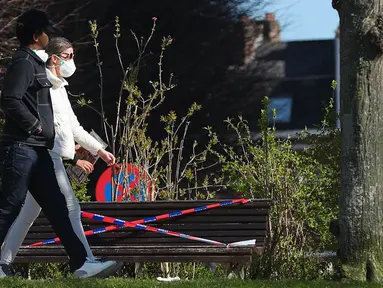 Orang-orang berjalan melewati bangku yang dipasangi garis polisi di Brussel, Belgia, pada 25 Maret 2020. Deretan bangku di sejumlah taman dilarang diduduki untuk mencegah orang-orang berkumpul atau berada terlalu lama di area publik tersebut. (Xinhua/Zheng Huansong)