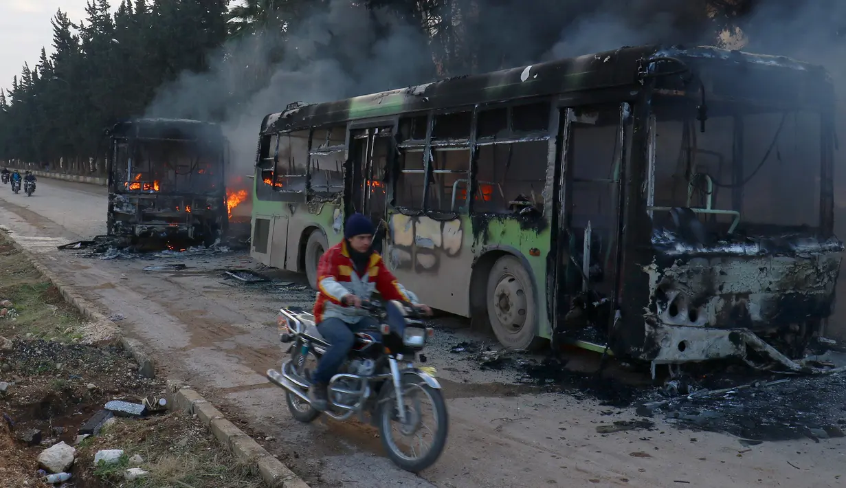Pengendara motor melewati bangkai bus yang dibakar di dekat Idlib, Suriah, Minggu (18/12). Sedikitnya lima bus yang sedang dalam perjalanan mengevakuasi warga sipil korban perang di Aleppo timur dibakar sejumlah orang bersenjata. (REUTERS/Ammar Abdullah)
