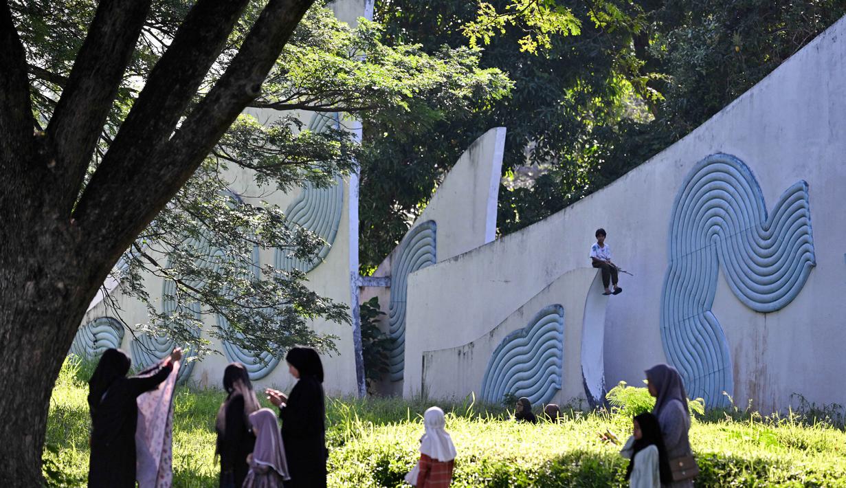 Momen ini menjadi refleksi untuk menyatukan kenangan pahit masa lalu dengan kepedihan akibat bencana alam terkini. Tampak dalam foto, seorang anak duduk di monumen peringatan sementara orang-orang berdoa untuk kerabat yang kehilangan nyawa dalam peristiwa gempa dan tsunami Aceh pada 2004 lalu di sebuah kuburan massal di Siron, pada 26 Desember 2025. (CHAIDEER MAHYUDDIN/AFP)