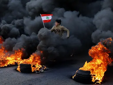 Demonstran antipemerintah melompati ban yang terbakar saat protes menentang elite penguasa di Kota Jal el-Dib, Lebanon, Selasa (14/1/2020). Demonstran menganggap elite penguasa gagal mengatasi ekonomi yang menurun tajam. (AP Photo/Bilal Hussein)