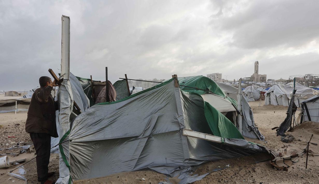 Ribuan tenda yang menjadi tempat tinggal keluarga pengungsi di Jalur Gaza rusak parah tersapu angin akibat badai musim dingin yang kuat. Tampak dalam foto, seorang pria Palestina yang mengungsi memperbaiki tenda yang didirikan di sepanjang pantai Kota Gaza saat angin musim dingin yang kencang menerpa wilayah tersebut pada Selasa 13 Januari 2026. (Omar AL-QATTAA/AFP)