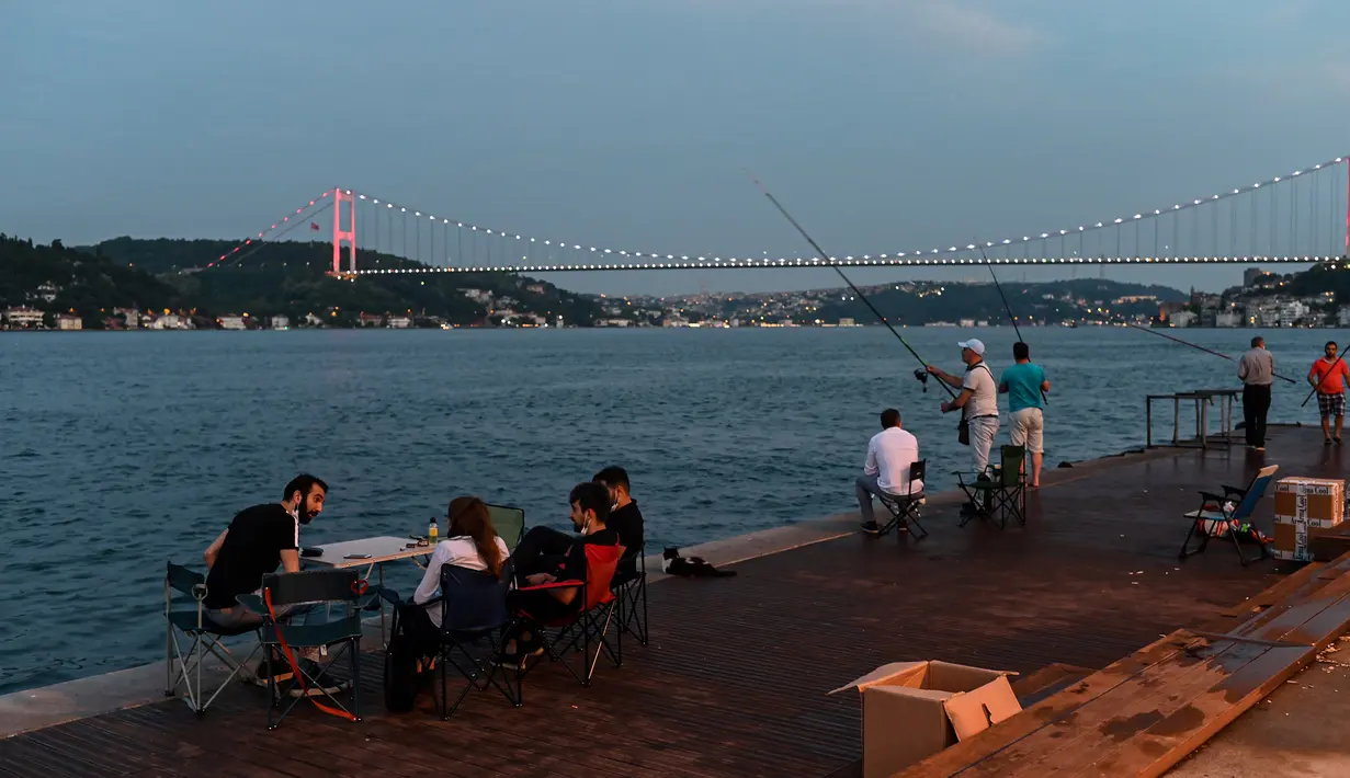 Orang-orang menikmati malam di sebelah pantai Bosphorus ketika jembatan Fatih Sultan Mehmet terlihat di Istanbul (6/7/2020). Ketika diselesaikan pada 1988, jembatan tersebut menjadi jembatan gantung terpanjang ke-5 di dunia; sekarang jembatan tersebut berada pada urutan ke-19. (AFP/Ozan Kose)