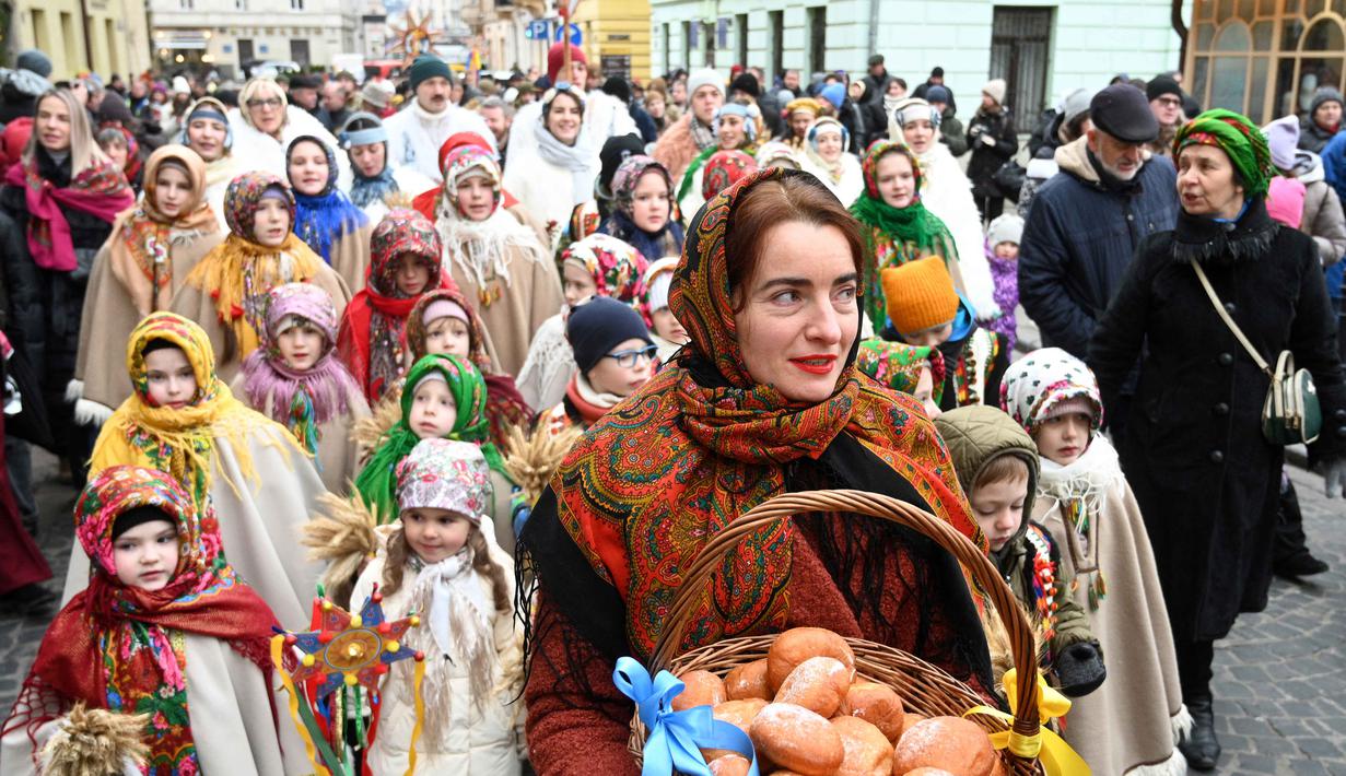 'Didukh' ditempatkan di sudut ruangan (biasanya di bawah ikon suci) selama Malam Natal. Tampak dalam foto, warga Ukraina yang mengenakan pakaian tradisional dan membawa hiasan Natal tradisional Ukraina 'Didukh' menyanyikan lagu-lagu rohani saat mereka mengikuti prosesi Malam Natal di Lviv pada 24 Desember 2025. (YURIY DYACHYSHYN/AFP)