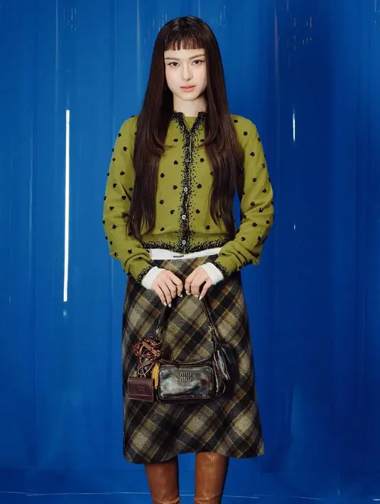 Prim Chanikarn, artis Thailand tampil dengan atasan model cardigan warna hijau motif hitam dipadukan rok tartan dan boots coklatnya. Ia membawa tas coklat.  [Dok. MiuMiu]