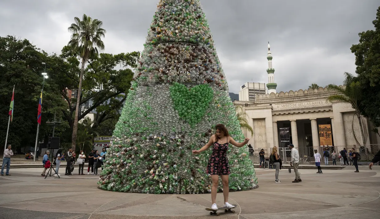 Seorang skaterboarder melewati depan pohon Natal yang terbuat dari botol plastik daur ulang di Caracas, Venezuela, Selasa (13/12/2022). Pohon Natal setinggi 10 meter tersebut merupakan proyek yang didukung kantor wali kota Caracas dan masyarakat umum yang berkontribusi dengan mengumpulkan botol dari jalanan. (Yuri CORTEZ / AFP)