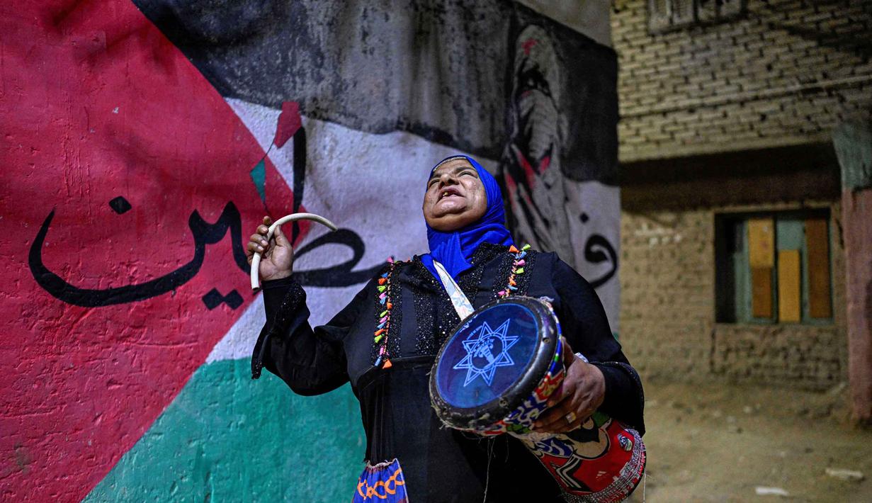Musaharati merupakan sebutan untuk petugas yang berkeliling permukiman menabuh genderang atau drum untuk membangunkan warga sahur saat bulan suci Ramadan. Tampa dalam foto, Dalal, seorang wanita Mesir 'musaharati', berdiri di depan grafiti yang menggambarkan bendera Palestina di sepanjang jalan sambil memukul gendang dan menyerukan umat Muslim untuk bangun dan makan sahur selama bulan suci Ramadan di distrik Hadayek el-Maadi, selatan Kairo pada Minggu 1 Maret 2026. (Khaled DESOUKI/AFP)