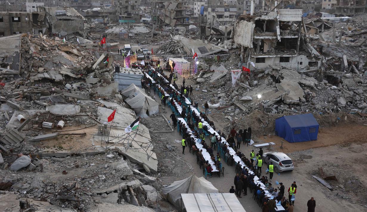 Lentera Ramadan kecil dan lampu hias yang terpasang di antara reruntuhan bangunan dan tumpukan puing, membawa sukacita dan kelegaan saat waktu berbuka puasa tiba. Tampak dalam foto, warga Palestina berkumpul untuk berbuka puasa bersama yang diselenggarakan oleh Yayasan IHH Turki, di tengah reruntuhan bangunan yang hancur di Kota Gaza pada Rabu 18 Februari 2026. (Omar AL-QATTAA/AFP)