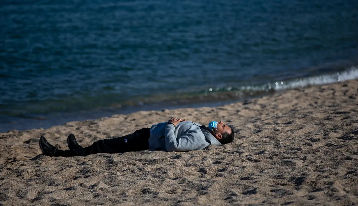 Seorang pria mengenakan masker beristirahat ketika penguncian untuk memerangi penyebaran virus corona berlanjut di sebuah pantai di Badalona, dekat Barcelona, Spanyol, Selasa, (28/4/2020). (AP Photo/Emilio Morenatti)