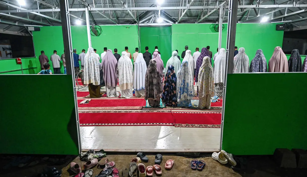 Diketahui, pemerintah setempat masih terus mengupayakan penyelesaian pembangunan Huntara. Tampak dalam foto, warga yang tinggal di tempat penampungan sementara bagi korban banjir melaksanakan salat berjamaah di sebuah aula di Meureudu, Kabupaten Pidie Jaya, Provinsi Aceh pada 21 Februari 2026, setelah banjir dan tanah longsor dahsyat melanda Sumatra, akhir November 2025 lalu. (CHAIDEER MAHYUDDIN/AFP)
