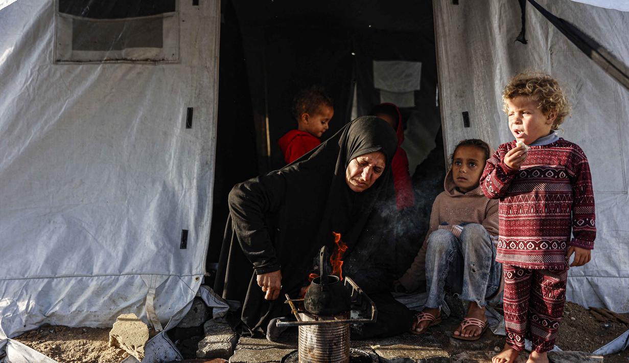 Philippe Lazzarini juga menekankan bahwa bantuan kemanusiaan yang diizinkan masuk saat ini jauh dari skala yang dibutuhkan untuk menangani krisis. Tampak dalam foto, seorang perempuan Palestina memanaskan panci sementara anak-anak memperhatikan di luar tenda tempat tinggalnya saat wilayah tersebut dilanda hujan dan kondisi musim dingin yang dingin, di Kota Gaza pada 28 Desember 2025. (Omar AL-QATTAA/AFP)