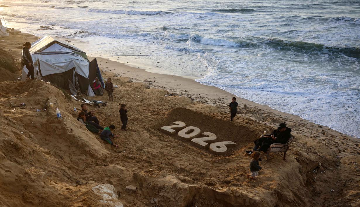 Sementara itu, sekitar 1,9 juta orang atau setidaknya 90% dari total populasi Gaza telah mengungsi setidaknya satu kali sejak perang meletus pada Oktober 2023 lalu. Tampak dalam foto, anak-anak berdiri di samping patung pasir karya seniman Yazed Abo Jarad (kanan) yang menggambarkan tahun mendatang di Deir El-Balah, Jalur Gaza tengah pada 30 Desember 2025. (Bashar Taleb/AFP)
