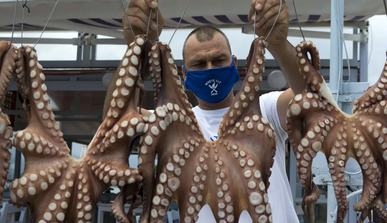 Pemilik restoran mengenakan masker menjemur gurita di Pulau Poros,  Yunani (7/8/2020). Terus bertambahnya jumlah kasus terkonfirmasi coronavirus di beberapa wilayah Yunani memaksa pihak berwenang memberlakukan jam malam pertama selama musim pariwisata di Pulau Poros. (Xinhua/Marios Lolos)