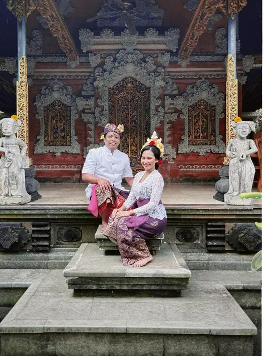 Indah memadukan kebaya Bali warna putihnya dengan kain kamen dan selendang warna ungu. Sedangkan Arie mengenakan baju safari, kain kamen, dan udeng. [Instagram/ariekriting]