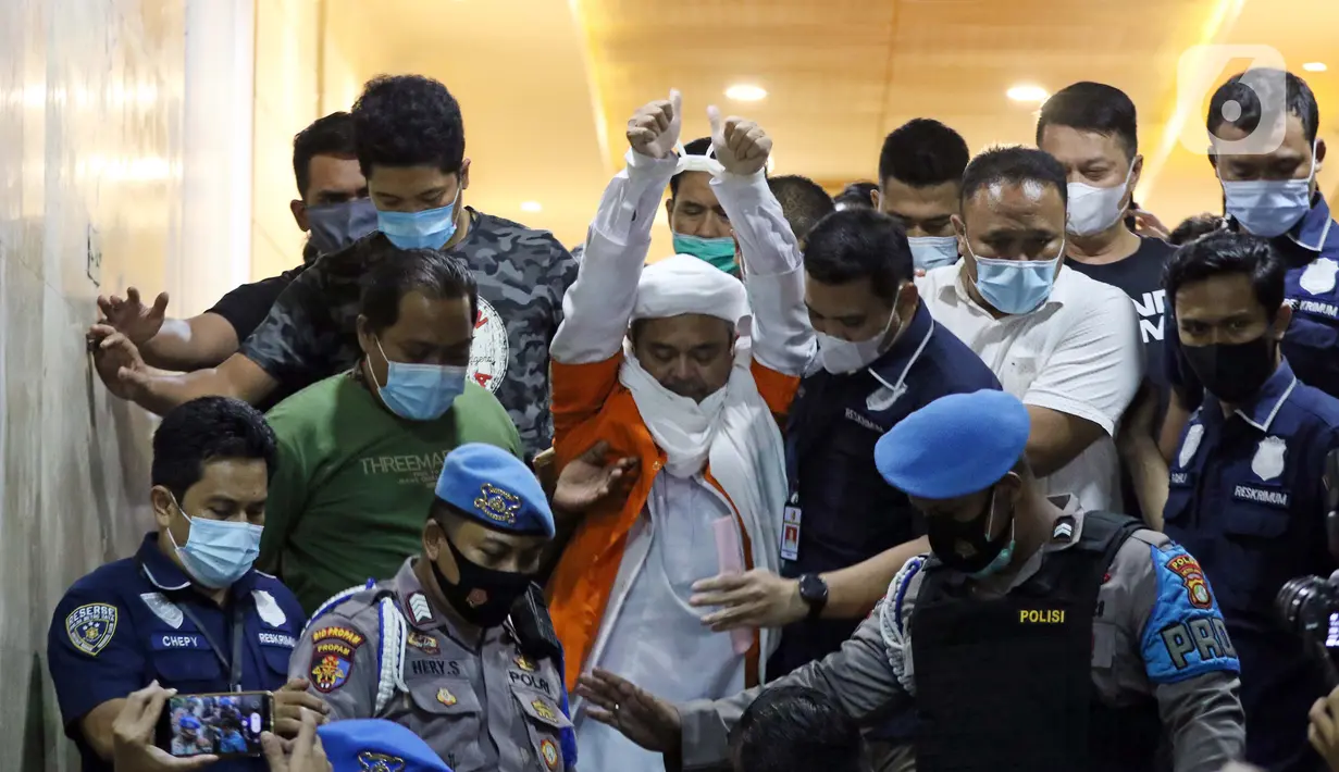 M Rizieq Shihab (tengah) mengangkat tangan saat meninggalkan gedung Ditreskrimum Polda Metro Jaya, Jakarta, Minggu dini hari (13/12/2020). Rizieq Shihab ditahan setelah menjalani pemeriksaan sebagai tersangka penghasutan dan kerumunan di tengah pandemi COVID-19. (Liputan6.com/Helmi Fithriansyah)