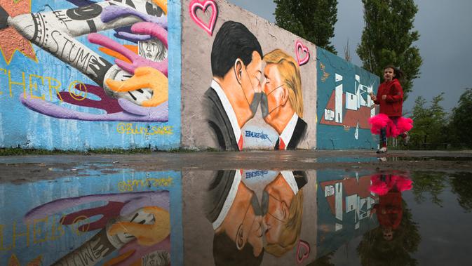 Seorang gadis berlari di depan Graffiti yang menggambarkan Presiden AS Trump (kanan) dan Presiden Tiongkok Xi Jinping saling berciuman dengan mengenakan masker di tembok taman umum Mauerpark di Berlin, Jerman, Rabu, (29/4/2020). (AP/Markus Schreiber)