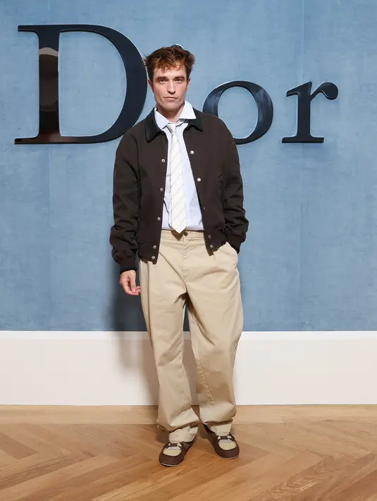 Robert Pattinson kenakan jaket bomber wol coklat Dior, kemeja katun bergaris, dan dasi sutra. Dipadu celana panjang lipit krem, dan sepasang sepatu kets suede coklat dan coklat. [Dior]