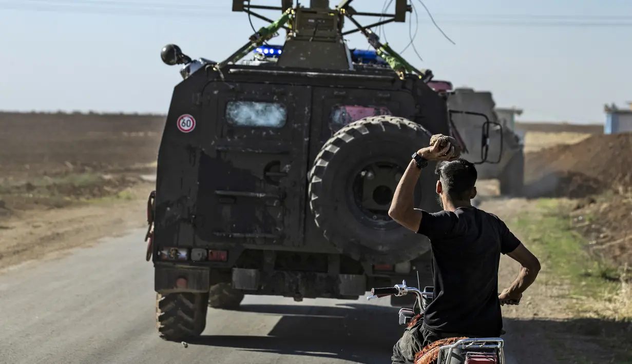 Seorang pria Kurdi menaiki motor melempar batu ke kendaraan militer Turki di dekat kota Al-Muabbadah, bagian timur laut Hassakah, Suriah (8/11/2019). Aksi dilakukan memprotes  terhadap serangan militer yang dilancarkan Turki. (AFP/Delil Souleiman)