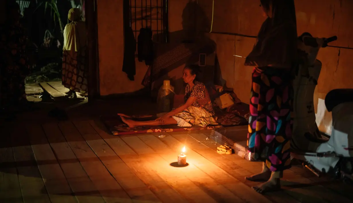 Hingga Selasa 16 Desember 2025 malam, warga yang mengungsi akibat banjir bandang pada akhir November 2025 bertahan hidup dalam kegelapan malam tanpa penerangan lampu listrik. Tampak dalam foto, Nurlimah (C), 65 tahun, yang tidak dapat berjalan sejak mengalami cedera dalam kecelakaan tabrak lari setahun lalu, duduk sambil berlindung di rumah tetangga yang sebagian runtuh di sebelah rumahnya, yang hanyut tersapu banjir bandang di Aceh Tamiang, pada Selasa 16 Desember 2025. (Yasuyoshi Chiba/AFP)
