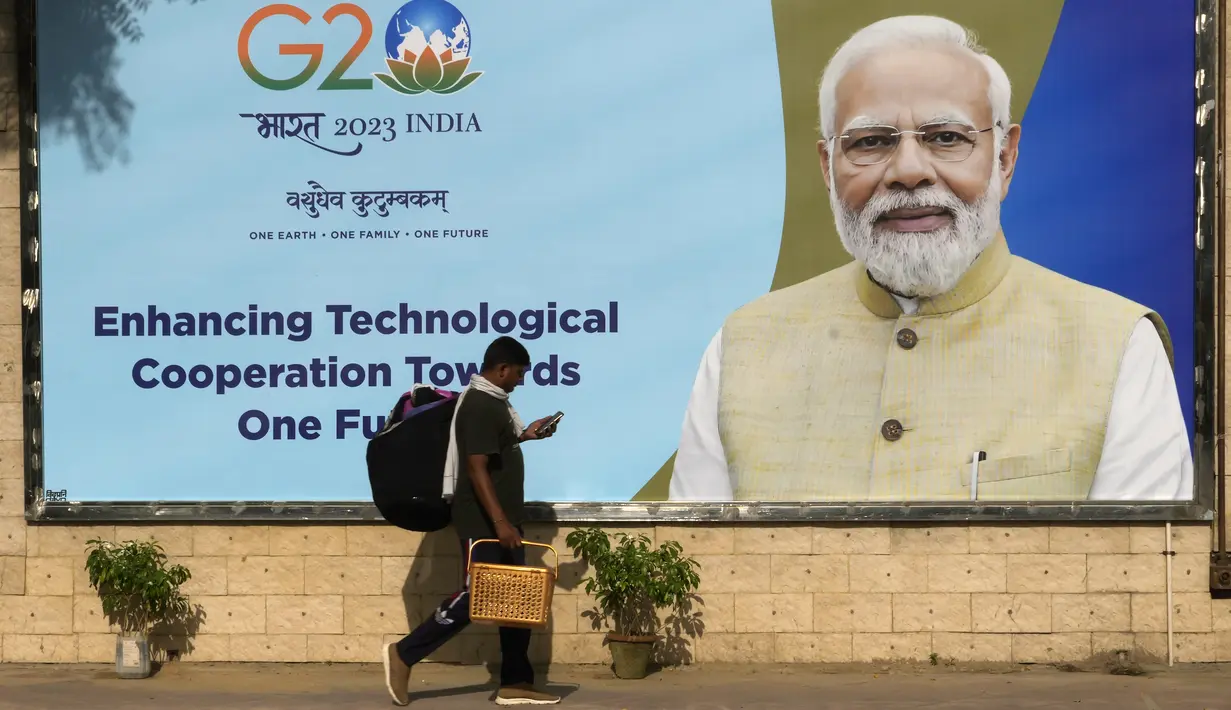 Seorang buruh migran memeriksa ponselnya saat ia berjalan melewati papan reklame bergambar Perdana Menteri India Narendra Modi, yang dipasang menjelang KTT negara-negara Kelompok 20 minggu ini di New Delhi, Senin, 4 September 2023. (AP Photo/Manish Swarup)