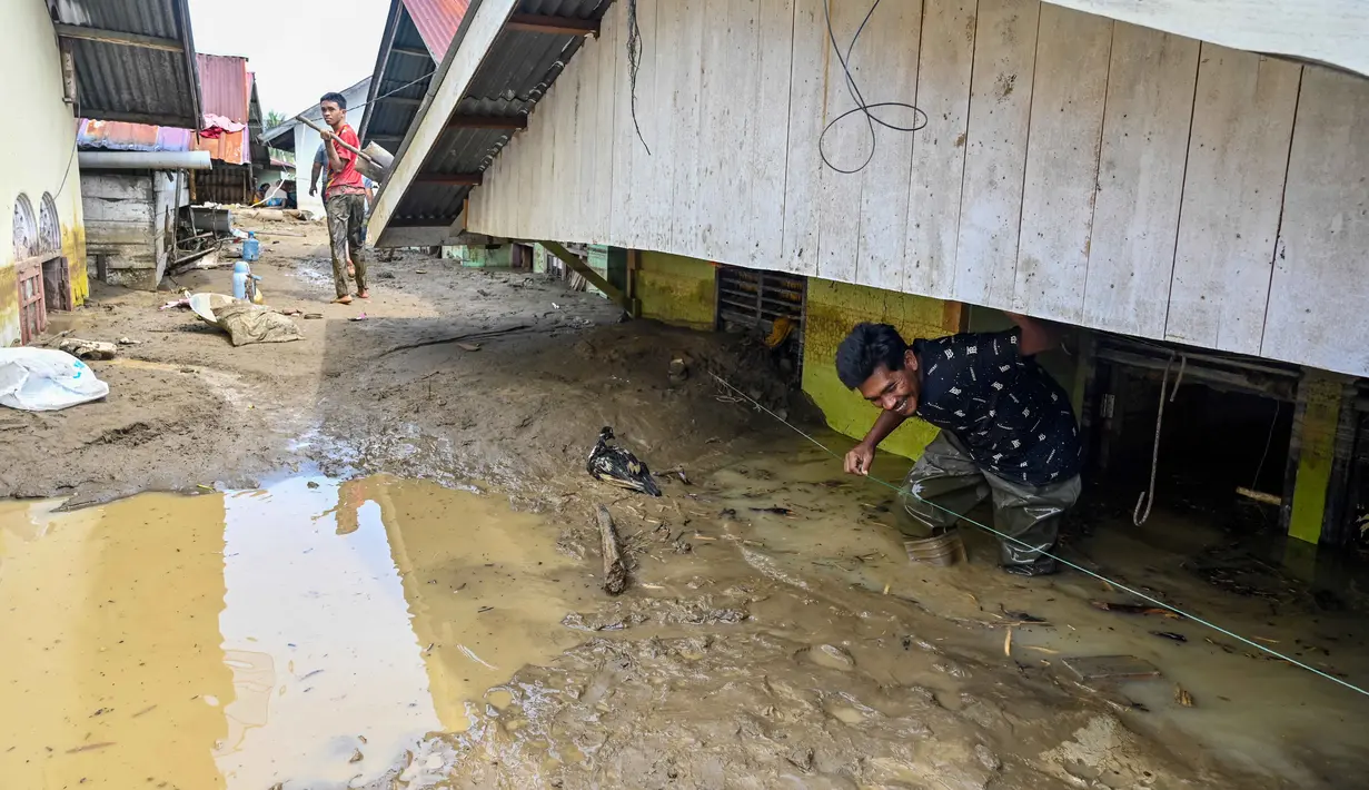 Warga berjalan melewati lumpur tebal di samping rumah-rumah yang terendam banjir bandang di Meureudu, Kabupaten Pidie Jaya, Provinsi Aceh, pada Jumat 28 November 2025. Permukiman warga di Meureudu, Kabupaten Pidie Jaya, Provinsi Aceh, terendam lumpur bercampur pasir sisa luapan banjir bandang yang melanda wilayah tersebut, sejak Selasa 25 November 2025. (CHAIDEER MAHYUDDIN/AFP)