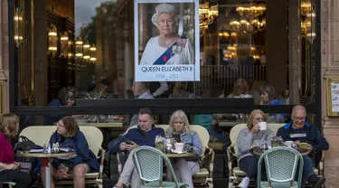 Orang-orang duduk di bar teras di sebelah potret Ratu Elizabeth II di pusat kota London pada 13 September 2022. Hotel, restoran, dan toko-toko penuh sesak saat penggemar kerajaan berdatangan ke jantung kota London untuk melihat jalan-jalan yang dipagari bendera, prosesi yang dipenuhi kemegahan, dan berani mengantre sejauh bermil-mil untuk kesempatan sekali seumur hidup mengucapkan selamat tinggal kepada Ratu Elizabeth II. (AP Photo/Emilio Morenatti, File)