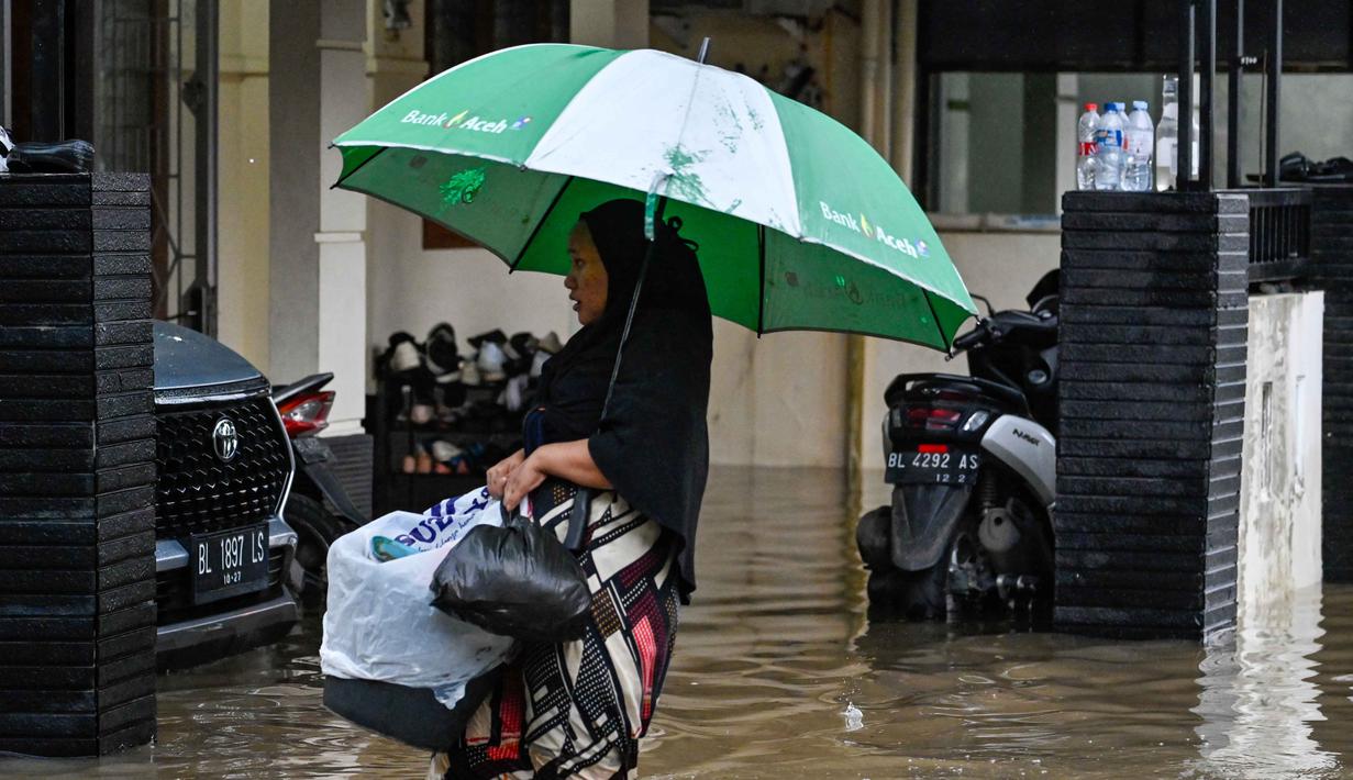 Banjir juga mengakibatkan 46 ribu rumah warga terendam air. Hingga kini, 1.497 jiwa harus mengungsi ke tempat yang lebih tinggi. Tampak dalam foto, seorang perempuan membawa barang-barangnya saat mengarungi banjir di jalan yang tergenang akibat hujan deras di permukiman Darul Imarah, pinggiran Banda Aceh, pada Kamis 27 November 2025. (CHAIDEER MAHYUDDIN/AFP)