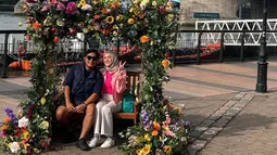 Sebelum masuk dan naik ke Inside Tower Bridge, Nina Zatulini dan sang suami, Chandra Tauphan Ansar berpose dulu di sebuah kursi kayu. Kursi yang berada di area wisata London Bridge itu dikelilingi berbagai macam bunga indah. Pasangan suami istri ini pun terlihat begitu ceria. (Liputan6.com/IG/@ninazatulini22)