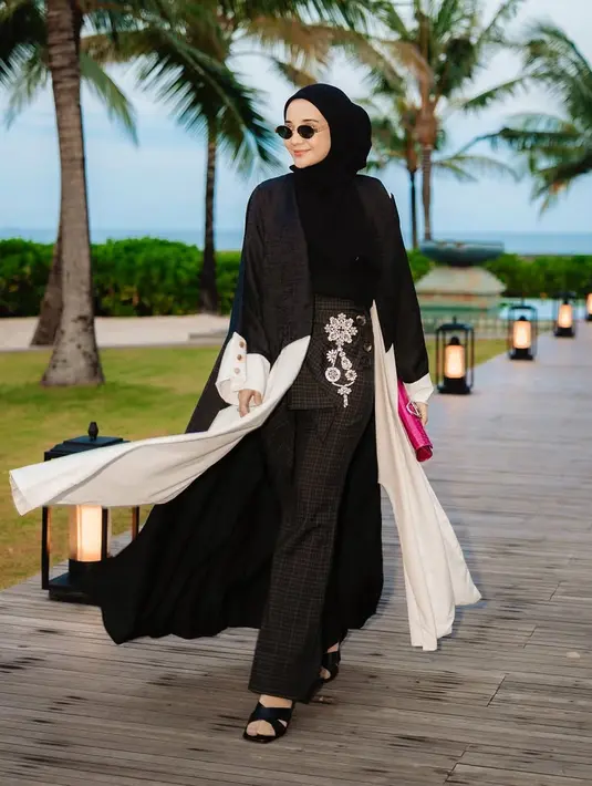 Zaskia Sungkar mengenakan outer panjang hitam dengan aksen putih di bagian bawah dipadukan inner dan kerudung warna senada. Serta bawahan celana panjang hitam bergaris dengan aksen apron dan bordiran bunga putih. [@zaskiasungkar15]