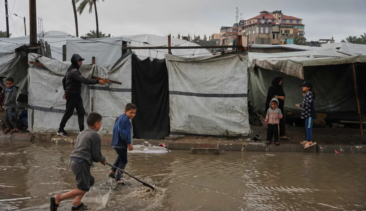 Air terlihat menggenangi jalanan dan memasuki tenda. Tampak dalam foto, anak-anak pengungsi Palestina berjalan melewati kamp tenda yang basah kuyup akibat hujan lebat di Deir al-Balah, Jalur Gaza tengah, Kamis, 26 Maret 2026. (AP Photo/Abdel Kareem Hana)