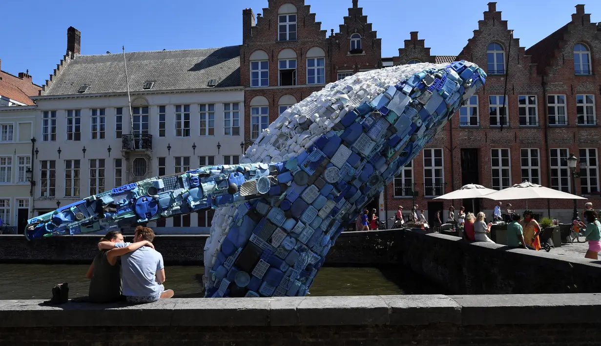 Wisatawan duduk dekat patung paus dari 5 ton sampah plastik yang ditampilkan di sungai Brugges, Belgia, Sabtu (14/7). Patung paus tersebut bertujuan untuk menyoroti ancaman yang diakibatkan oleh penggunaan plastik secara besar-besaran. (AFP/JOHN THYS)